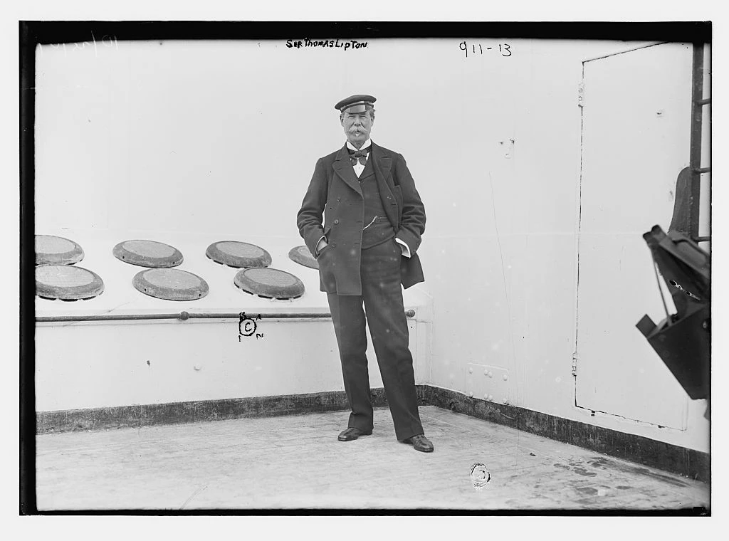 Thomas Lipton standing on a ship’s deck in a cap and dark suit; complete black-and-white archival frame with glass-negative border and handwritten identification.