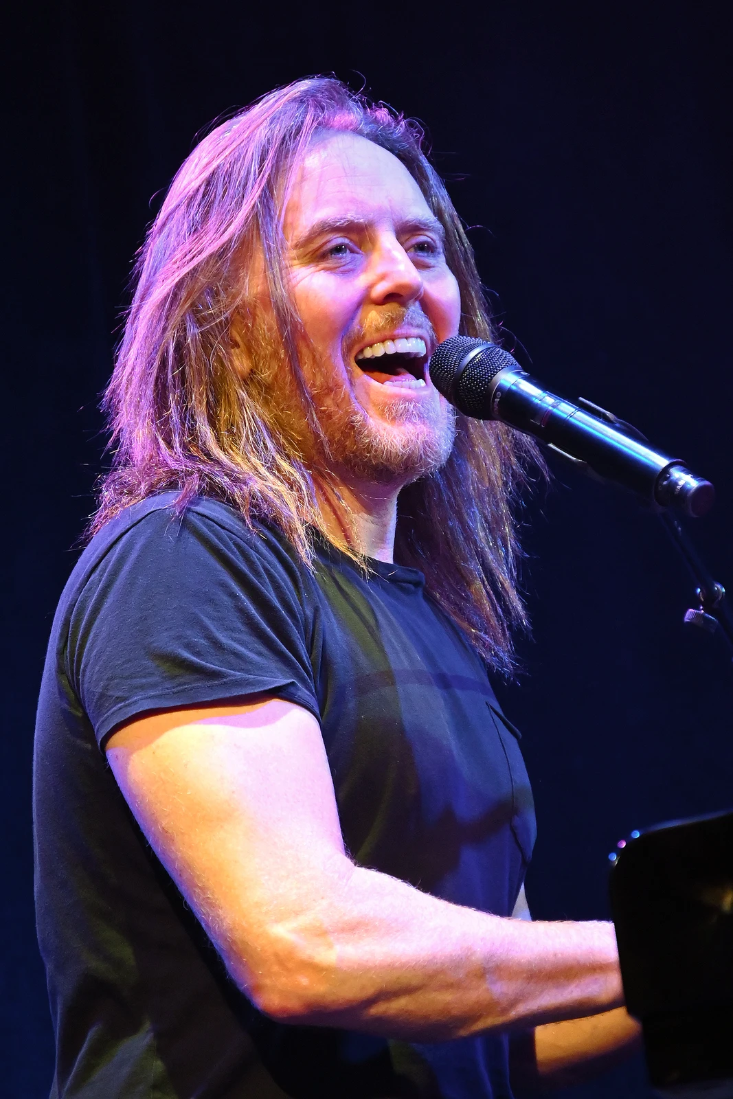 Tim Minchin performing at a microphone at Town Hall in New York, with the complete published portrait, arm, piano edge and dark background retained.