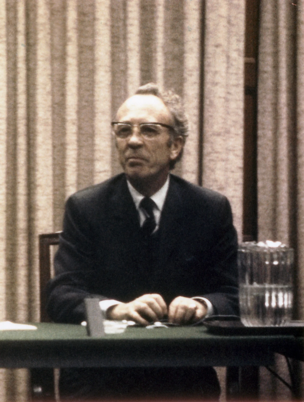 Tommy Douglas seated behind a table with a microphone and water jug in a complete supplied photograph dated circa 1971.