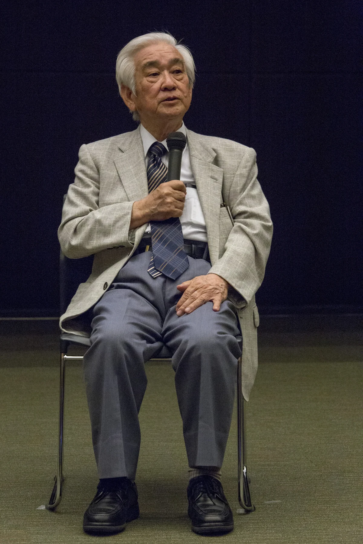 Toshihide Maskawa seated and holding a microphone in OIST’s complete supplied photograph.