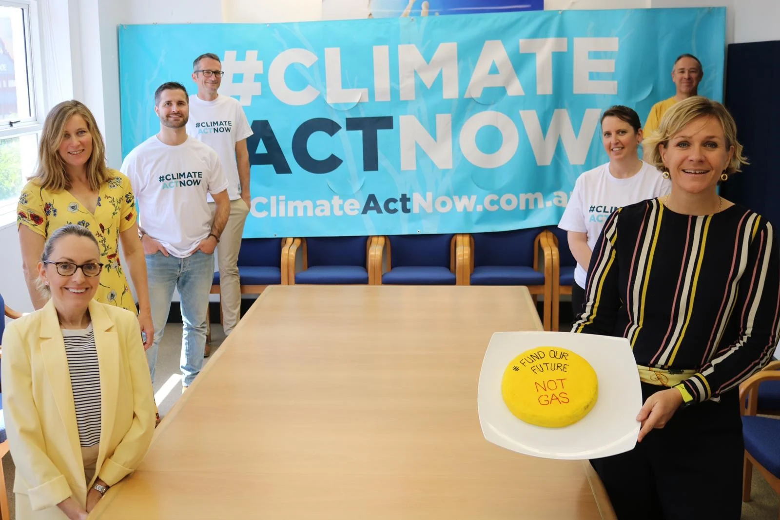 Zali Steggall at right holding a cake in her electorate office with climate activists around a table and a climate-action banner behind them.