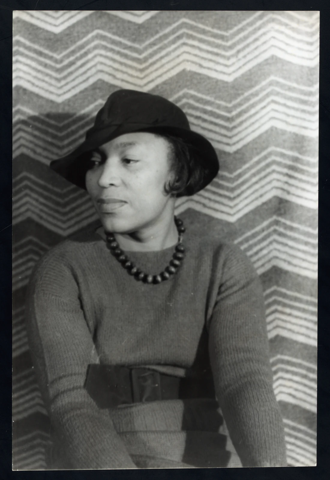 Zora Neale Hurston in a hat and large beaded necklace, photographed against a patterned backdrop by Carl Van Vechten in 1938